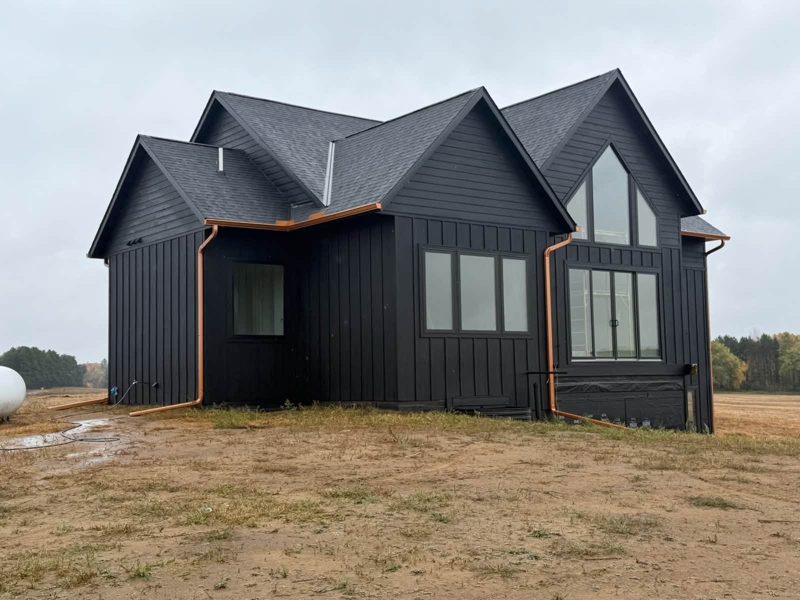 Black board-and-batten house with copper-colored gutters and downspouts at the corners